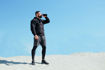 pumped up athlete drinks water and rests after jogging on a sandy mountain