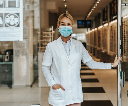 Young And Attractive Female Optometrist With Face Protective Mask Standing At Open Optical Store Doors And Looking Outside. She Is Confident And Serious. Covid-19 Open For Business Concept.