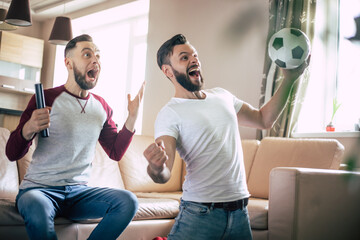 Two best friends and fans of football watching some sport match on the TV and drinking beers and...