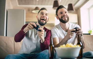 Excited smiling men playing in video games on tv at home on the couch. Friends with joysticks play...