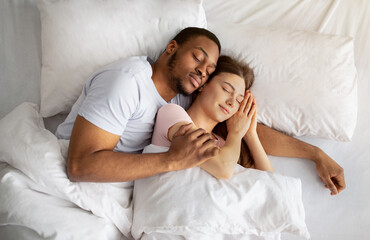 Loving multiracial couple having cozy morning, cuddling together in their sleep on bed, top view