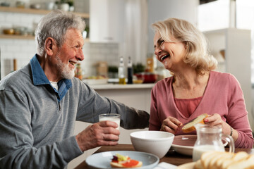 Senior couple eating breakfast in the kitchen. Husband and wife talking and laughing while eating a sandwich..