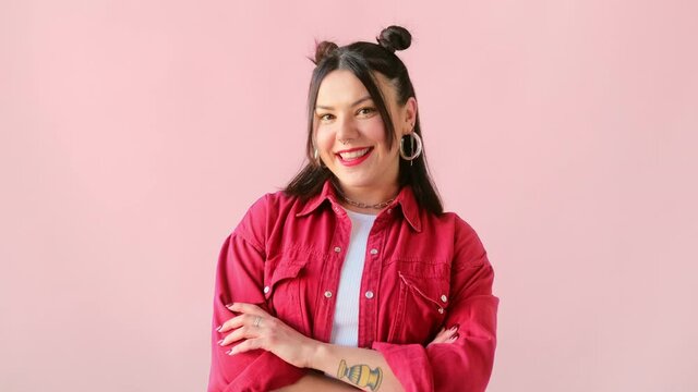 Stylish brunette woman with tattoo blinking eye, smiling and looking at camera on pink background. Winking and flirting trendy female.