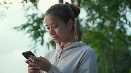 Asian sport woman typing on mobile phone with smile in the public park - Powered by Adobe