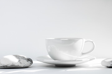White tea or coffee mug and saucer, on a white table in the sun, with harsh shadows, and stones. Creative solution poster with a drink. Copy space