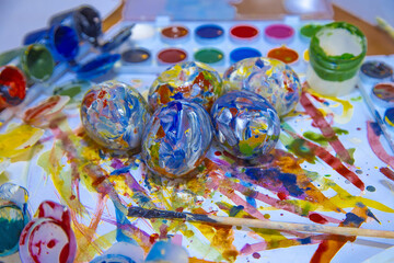 Fototapeta premium Easter colorful eggs lie on a brightly painted white background, surrounded by paints and brushes for resovaniya. Bright Easter eggs, side view.