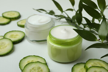 Jars with cosmetic cream, cucumber slices and branch on white table