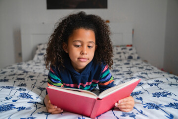 Afro little girl laying on bed with a red book reading front view