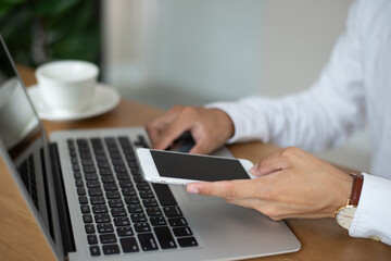 Young businessman hand holding smartphone and checking and replying to emails in the laptop on office desk.