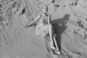 Fototapeta na wymiar Full length portrait. Beautiful blonde woman posing in embroidery dress on desert, lying on sand. Black and white photo.