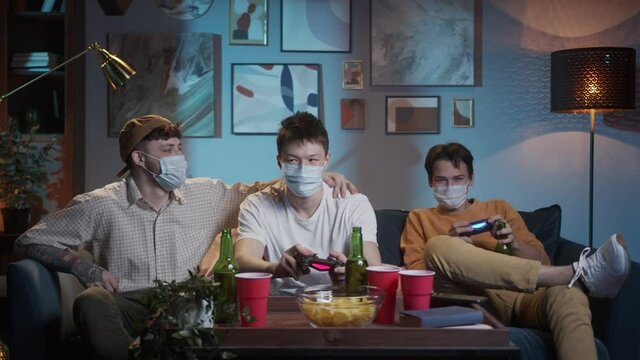 Millennial international friends wearing medical protective masks playing video games, gaming together at home. Young people having fun with new trends technologies during coronavirus covid 19. 