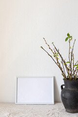 Home decor mock-up, blank picture frame near white painted concrete wall , black earthenware jug with pussy willow branches and blossoming green leaves