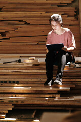 Young female carpenter working on design of furniture item for customer and drawing sketch she sitting on pile of wooden planks