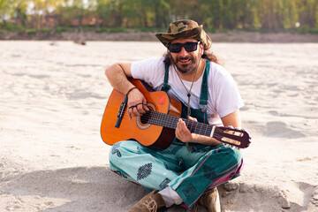 Hippy man with hat beard and glasses playing guitar outdoors on the beach