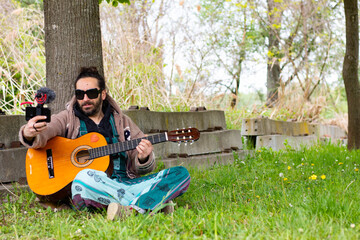 Hippy man with beard and glasses playing guitar making a video herself outdoors on the grass