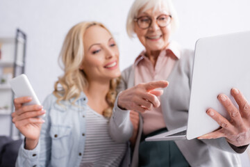 Senior woman on blurred background pointing at laptop near daughter with smartphone