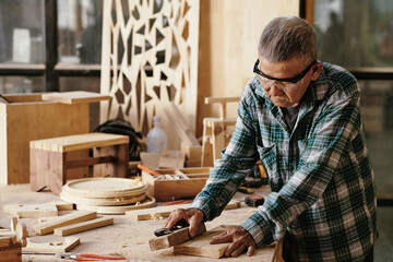 Senior carpenter polishing small board with wooden block at his workbench