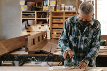 Elderly experienced carpenter in protective mask polising wooden plank with electric sander