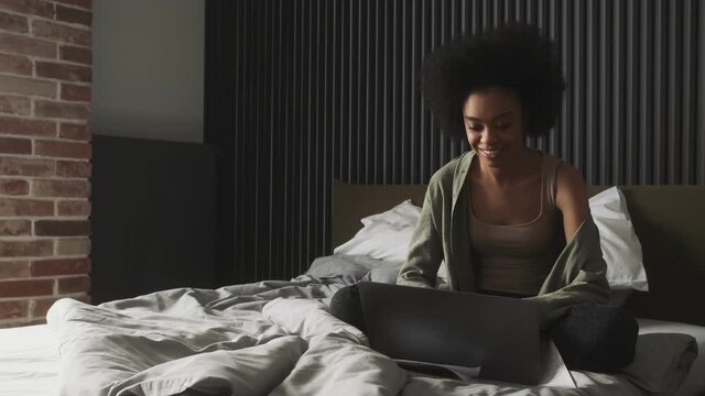 The black smiling woman sitting on the bed with laptop and looking at his screen in the room