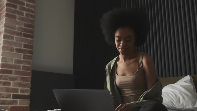 A black happy woman sitting on the bed in front of laptop and typing something on keyboard in the room