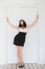 sexy brunette in a short dress stands near white doors