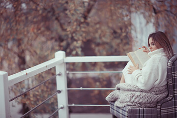 Fototapeta premium girl in a knitted sweater reads a book on the veranda in the park, seasonal romance concept