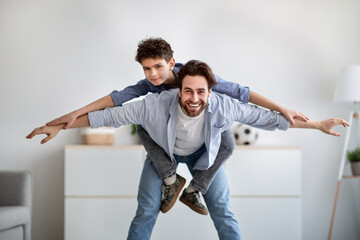 Playful father and son having fun together, daddy riding boy on back like flying on plane at home in living room