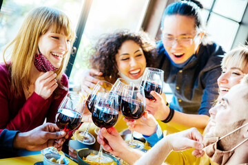 Happy friends wearing protective face masks toasting red wine sitting at bar restaurant - New normal friendship concept with young people having fun at home party - Focus on glasses	
