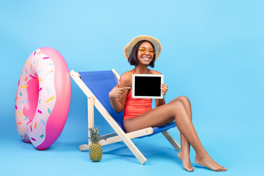 Black woman in swimsuit pointing at tablet pc with blank screen while sitting in lounge chair on blue background, mockup - Powered by Adobe