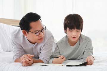 Asian father teaching homework to his son in bed for homeschooling and education concept