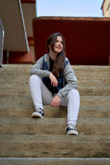 long-haired, fair-skinned teenager in denim jacket and white pants sitting on white stairs. Girl in urban style smiling and pensive. empowered young woman on the street