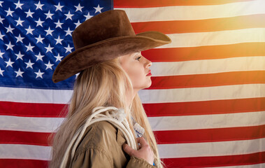 A blonde patriotic lady in a cowboy hat and with a lasso stands against the background of the...