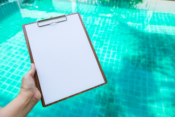 Black white paper on wooden clipboard over blurred blue water background, pool maintenance report, outdoor day light