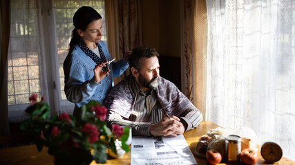 Portrait of poor woman cutting husband's hair indoors at home, poverty concept.