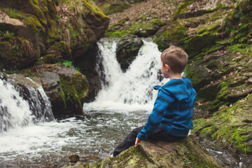 Naklejka na ściany i meble Kid sitting on the rock and looking at waterfall on the river in the rocks. Tourism with children concept.