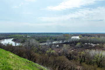 Fototapeta na wymiar Panoramic view from the high bank to the river, forest, green meadow and beautiful fields. View from the high bank to the spring river and the trees around.