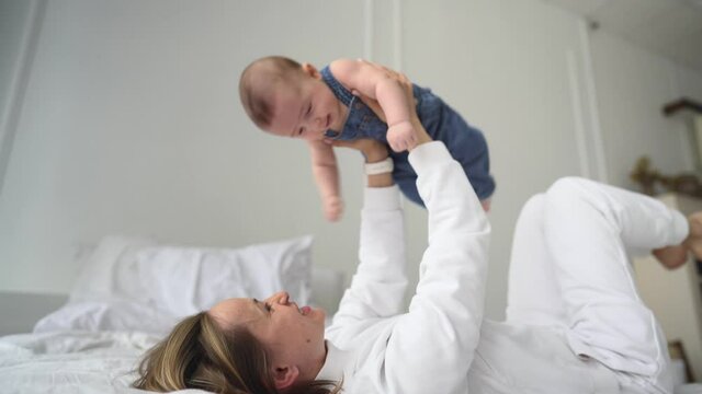 Happy healthy young adult caucasian mum lifting cute infant child up playing airplane on bed. Loving caucasian mom doing yoga gymnastic exercise with funny adorable baby son in cozy bedroom