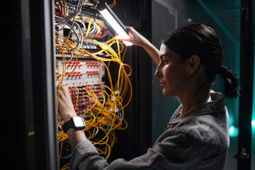 Side view portrait of female network engineer connecting cables in server cabinet while working with supercomputer in data center