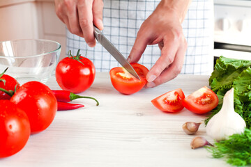 Man's hands cuts a ripe tomato with a knife on wooden white table. Process of making salad. Healthy food concept. Close-up.