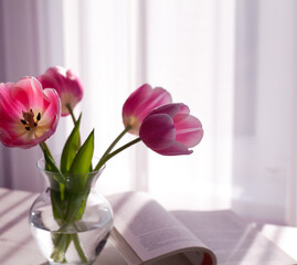 Bouquet of pink tulips in glass vase and next to it lies an open book. Selective focus.