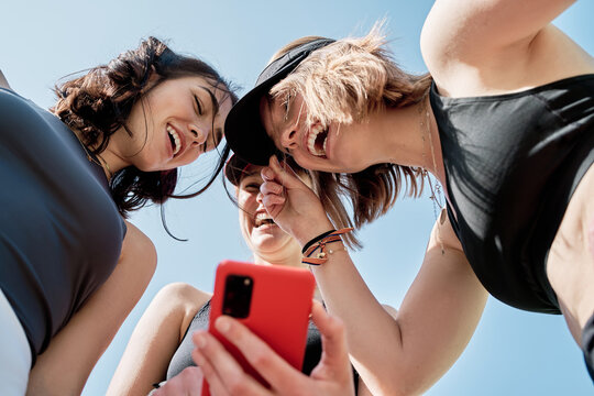 Three Beautiful Young Friends Laughing While Looking At Their Cell Phones After Practicing Sports,view From Below Of Three Smiling Girls With Their Cell Phones.