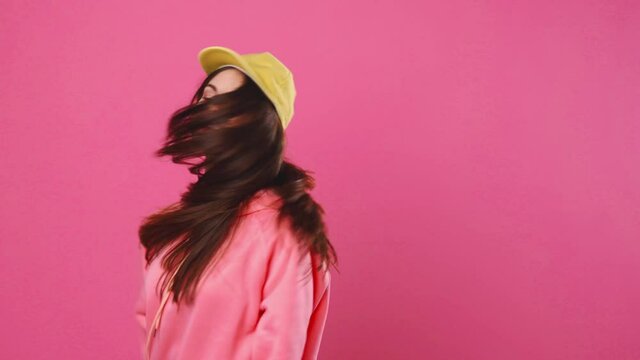Happy woman in yellow cap and pink sweatshirt jumping and laughing in front of the camera in the studio on a pink background