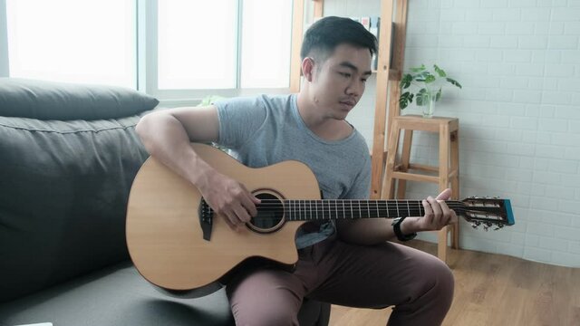 Musician Asian male playing acoustic guitar instrument singing song learning practicing carefree time spend activity occupation sitting on sofa in living room quarantine lockdown staying safe at home