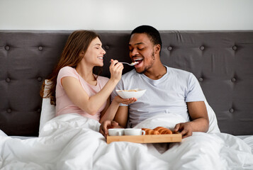 Happy multiracial couple having breakfast in bed, young woman feeding her husband with tasty cereal