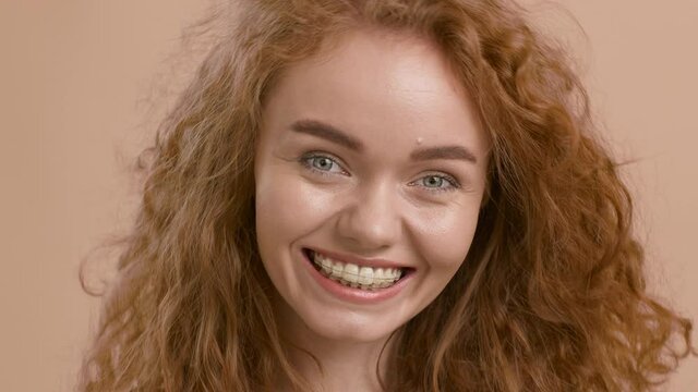 Red-Haired Woman With Ceramic Braces Smiling Posing Over Beige Background