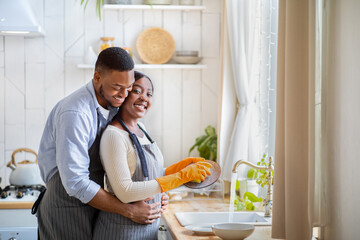 Handsome black man hugging his girlfriend while she is washing dishes at home, free space