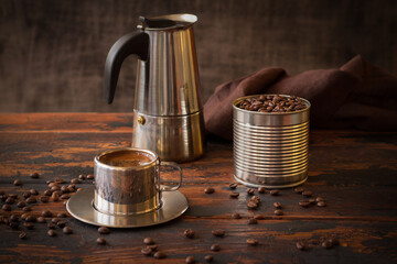 Steel cup of coffee, blended coffee beans in a tin can and geyzer coffee maker, brown napkin on a dark rustic wooden table among scattered coffee beans, blended, vintage