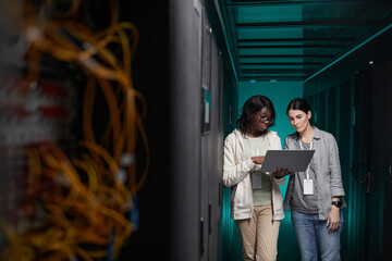 Wide angle portrait of two young women using laptop in server room while setting up supercomputer...