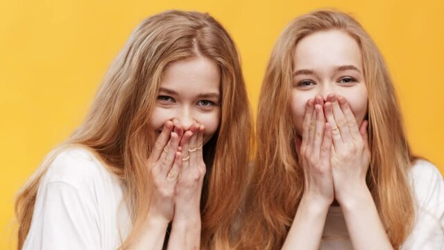 Shocking news. Close up portrait of surprised twin sisters covering mouths in amazement, laughing over orange background