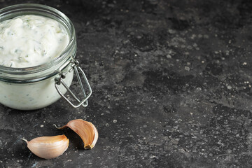 Greek tzatziki sauce in a glass jar with cloves of garlic on a dark background copy space